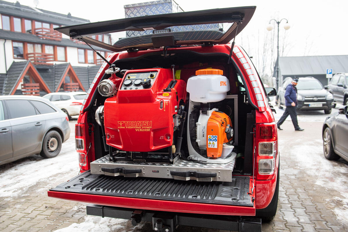 Zdjęcie pokazuje otwarty bagażnik samochodu strażackiego typu pickup. Widać stojacą w bagazniku dużą pompę do pobierania wody oraz inne urządzenia. Samochód stoi na parkingu, w tle inne auta